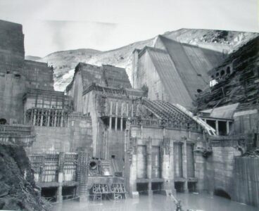 Fotografía antigua en blanco y negro que documenta el proceso de construcción de la presa y la central hidroeléctrica de Salime, perteneciente a Saltos del Navia en Asturias, con grandes estructuras de hormigón y andamios entre montañas.