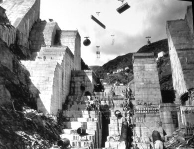 Fotografía histórica en blanco y negro de las obras de construcción de la presa de Salime en el río Navia, Asturias, mostrando los grandes bloques de hormigón y el sistema de cables para el transporte de materiales.