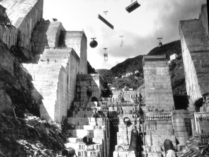 Fotografía histórica en blanco y negro de las obras de construcción de la presa de Salime en el río Navia, Asturias, mostrando los grandes bloques de hormigón y el sistema de cables para el transporte de materiales.