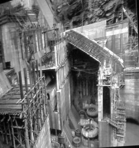 Fotografía histórica en blanco y negro que muestra los trabajos de construcción de la presa y la central de Salime, con andamios y piezas de turbinas en el lecho de la obra.