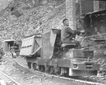 Fotografía histórica en blanco y negro de un trabajador operando una locomotora de vía estrecha con vagonetas durante las obras de construcción de la presa de Salime en el río Navia, Asturias.