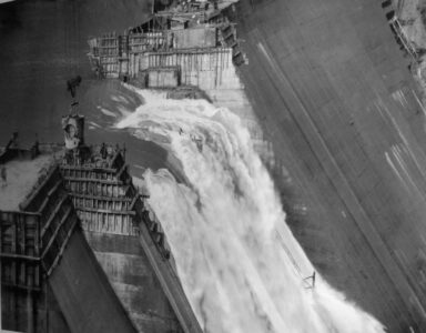 Fotografía histórica en blanco y negro de las obras de construcción de la presa de Salime, mostrando una gran caída de agua a través de la estructura de hormigón y los andamios en el río Navia.