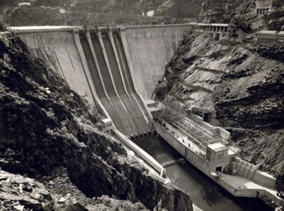 Fotografía histórica en blanco y negro de la construcción de la presa de Salime y su central hidroeléctrica en el río Navia, Asturias, destacando sus grandes aliviaderos y el entorno montañoso.