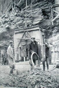 Fotografía histórica de obreros operando un gran cubilote de hormigón suspendido por cadenas durante las obras de construcción de la presa de Salime en los Saltos del Navia, Asturias.